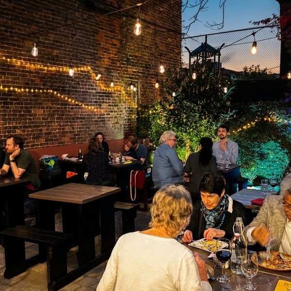 Patrons enjoying an evening on a brick-walled outdoor patio lit with string lights and colorful foliage.
