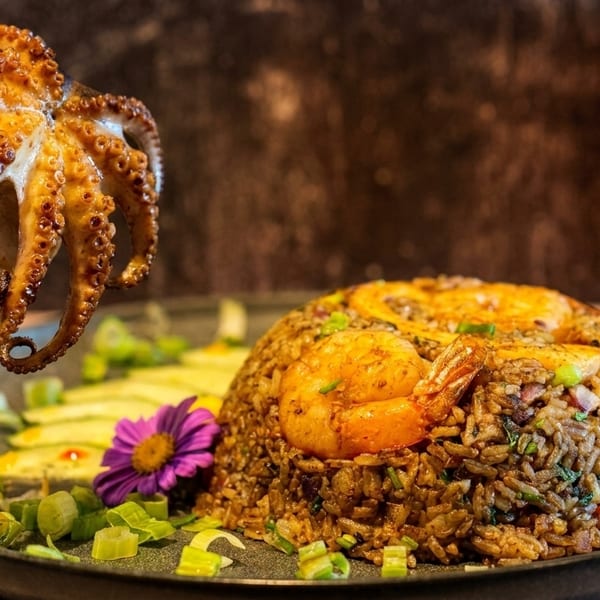 Grilled octopus on a fork above shrimp fried rice, garnished with scallions and a purple flower.